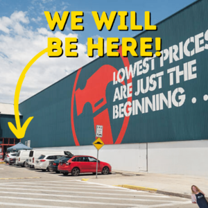 An image of a Bunnings Warehouse exterior with a large red and white Bunnings logo. A yellow arrow and text overlay in the same color state "WE WILL BE HERE!" indicating the location of the upcoming Bunnings Sausage Sizzle event in the parking area. The slogan "Lowest prices are just the beginning" is visible on the wall, setting the scene for a community gathering.